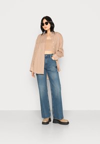 Camisa oversized às riscas beje e branca, top curto bege, jeans azuis de cintura alta e sapatos plataforma chunky bege. Óculos de sol redondos pretos.