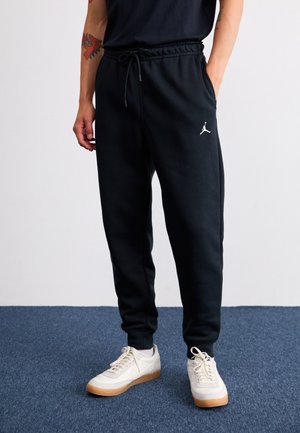 Mann trägt schwarze Jordan-Sweathosen mit weißen Sneakers, steht auf blauem Teppich, eine Hand in der Tasche und Drachen-Tattoo am Arm.