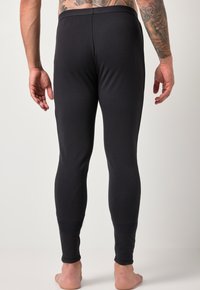 Hombre con tatuajes en la parte superior de la espalda y el brazo, vistiendo leggings negros ajustados, de pie descalzo contra un fondo claro y sencillo.