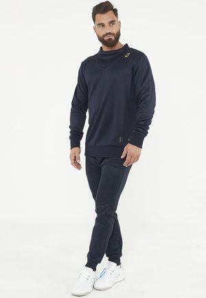 Man met baard die een marineblauw trainingspak en witte sneakers draagt, staat met gekruiste benen en kijkt opzij tegen een witte achtergrond.