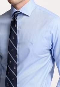 Camisa de vestir azul claro con diseño ajustado, combinada con una corbata azul marino con patrones de anclas y rayas diagonales. Detalle sutil de logo en la camisa.