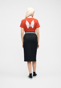 Γυναίκα με μακριά ξανθά μαλλιά που φορά κόκκινο μπλουζάκι με λευκό σχέδιο φτερών και παπουτσιών στην πλάτη, μαύρη μίντι φούστα και μαύρα ψηλά αθλητικά παπούτσια.