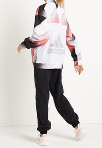 Adidas track jacket with a geometric red, black, and white pattern, featuring a mesh back and black joggers, worn with light pink sneakers.
