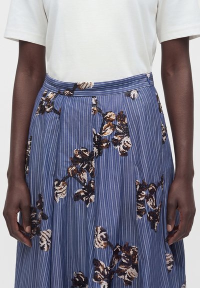 Jupe à fines rayures bleues avec broderie de sequins floraux foncés, portée avec un haut blanc uni à manches courtes, les mains reposant sur les côtés.