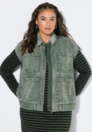 Kvinde med langt bølget hår iført øreringe, en grønstribet kjole og en ærmeløs vasket denimvest med store lommer, stående med hænderne på hofterne.