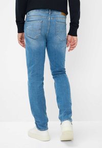 Jeans in denim blu chiaro con vestibilità slim, dotati di due tasche posteriori e passanti per la cintura. Indossati con sneakers bianche, evidenziando un design pulito.