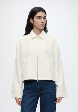 Frau mit langen dunklen Haaren, die eine weiße, strukturierte Reißverschlussjacke mit Kragen und dunkelblaue Jeans trägt, steht vor einem schlichten weißen Hintergrund.