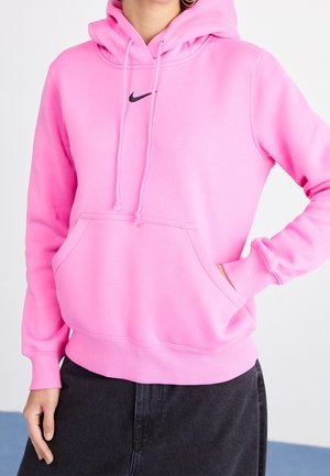 Person trägt einen knallpinken Nike-Hoodie mit Fronttasche und Kordeln, kombiniert mit dunklen Hosen, Hand teilweise in der Tasche.