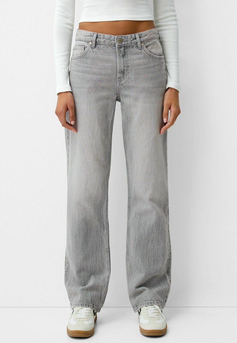 Personne portant un jean gris clair à jambes larges, un crop top blanc à manches longues et des baskets blanches avec semelles marron, debout devant un fond uni.