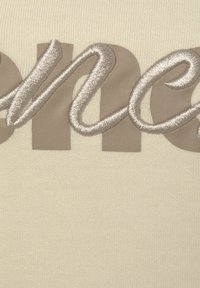 Crème-kleurige stof met grote, beige gestikte letters, met een gladde, licht gestructureerde oppervlakte en ingewikkelde borduurdetails.