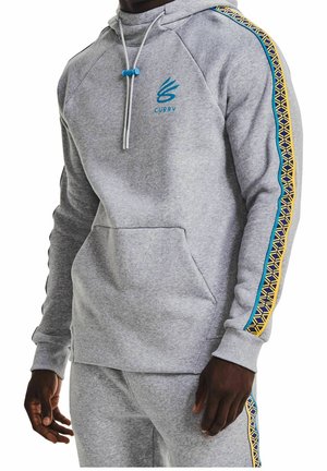 Homme portant un sweat à capuche gris clair et un pantalon de jogging assorti avec des rayures bleues et jaunes à motifs sur les manches et les jambes ainsi que le logo "Curry" sur la poitrine.