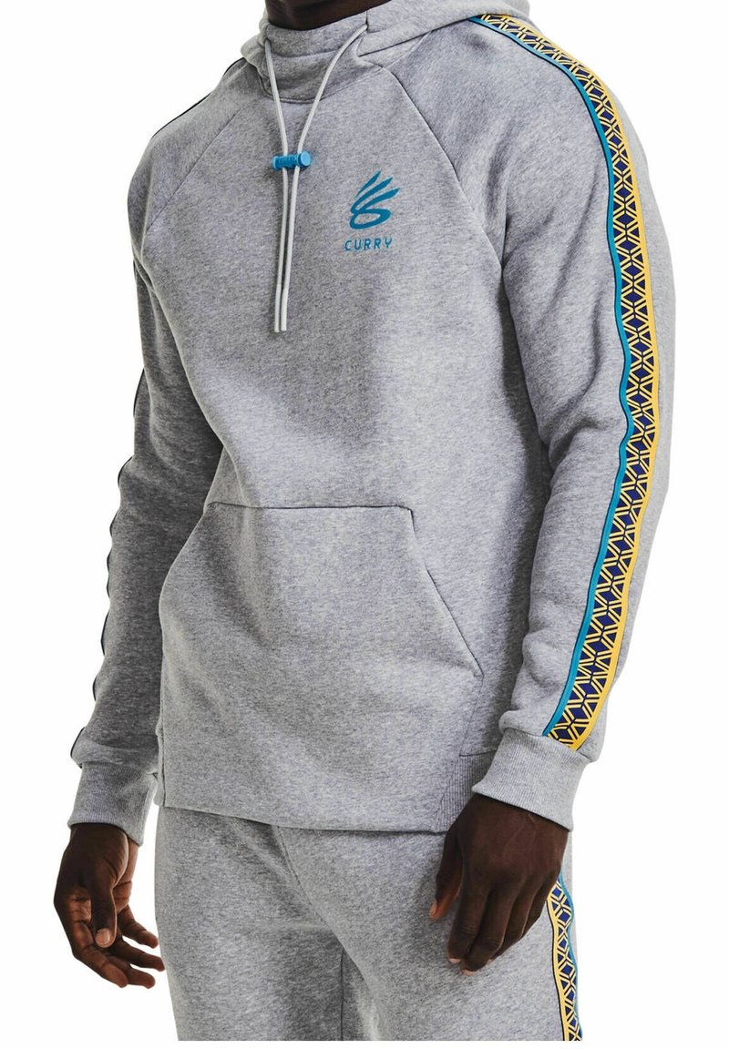 Homme portant un sweat à capuche gris clair et un pantalon de jogging assorti avec des rayures bleues et jaunes à motifs sur les manches et les jambes ainsi que le logo "Curry" sur la poitrine.