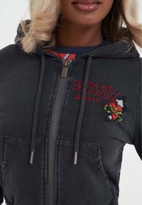 Černá mikina na zip s červeným textem "Ed Hardy designs" a vyšívaným srdcem s ostnatým drátem, kterou nosí osoba se světlými blond vlasy.