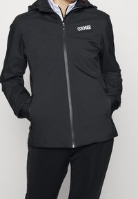Person mit schwarzer Colmar-Jacke mit Reißverschluss, die die Hände in den Taschen hat und schwarze Hosen trägt, vor einem schlichten hellen Hintergrund.