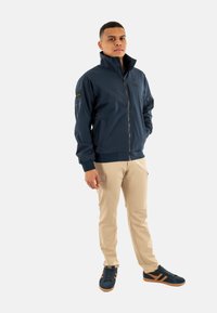 Chaqueta cortavientos azul marino con cuello alto, cremallera frontal y puños de canalé. Pantalones cargo beige y zapatillas azul marino con franjas naranjas.