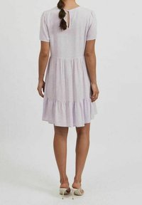 Žena v světlé fialové, krátkropřísné šaty po kolena s jemným tečkovaným vzorem a volánkovým lemem, viděná ze zad.