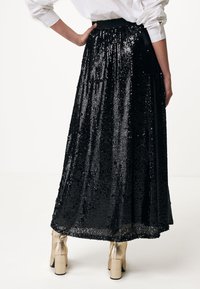 Schwarzer Pailletten-Maxikleid mit fließendem Design, hoher Taille und glänzender Textur. Kombiniert mit goldfarbenen Blockabsatzschuhen.