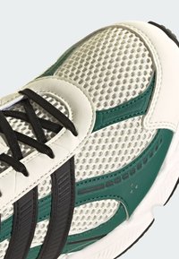 Tenisky s textilným vrchom v bielej farbe, s zelenými akcentmi a čiernymi pruhmi. Zakrivená špička, textúrovaný povrch a čierne šnúrky.