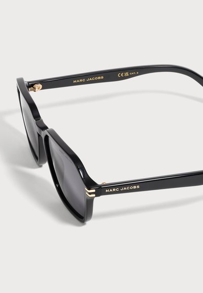 Lunettes de soleil noires Marc Jacobs avec des verres carrés foncés et un marquage doré sur les branches, exposées sur une surface blanche.