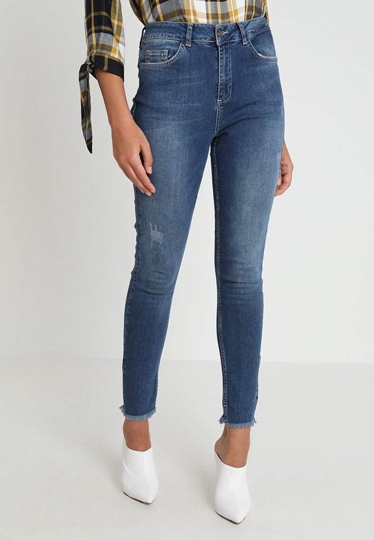 Jean en denim bleu taille haute avec une coupe ajustée, un léger effet usé et un ourlet brut, associés à une chemise à carreaux et des talons blancs.