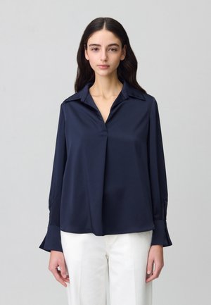 Blouse marine avec une texture douce et soyeuse, dotée d'un col classique, de manches longues et d'une coupe décontractée, associée à un pantalon blanc.