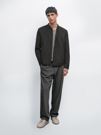 Mørkebrune uldblandingsjakke med ståkrave, knaplukning og sidelommer, båret over en lys grå skjorte og grå bukser, parret med beige sneakers.