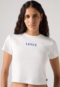 Maglietta bianca cropped realizzata in tessuto morbido con logo "LEVI'S" ricamato in blu sul petto e maniche corte. L'orlo termina sopra la vita.