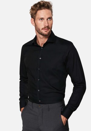 Black long-sleeve shirt in smooth cotton with a pointed collar, front buttons, and a tailored fit. Worn with gray tailored trousers.