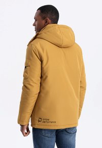 Gul fodrad jacka med huva, försedd med en sidficka på ärmen och en logotyp nära fållen, ihop med blå jeans.