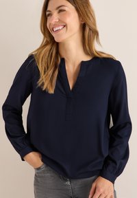 Blouse bleu marine en tissu lisse, avec un décolleté en V, des manches longues à poignets élastiques et une coupe décontractée. Portée avec un jean gris.