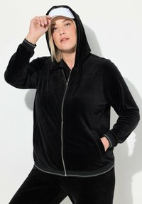 Sudadera con cremallera de terciopelo negro, con capucha, bolsillos frontales y puños acanalados en contraste. Modelo con una visera blanca. Textura suave.
