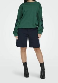 Sudadera verde de manga larga con bordado floral, combinada con pantalones cortos azul oscuro y botas negras hasta el tobillo. Textura de tejido suave.