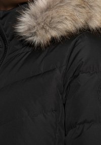 Chaqueta acolchada negra con un cuello alto, que presenta un ribete de pelaje sintético desmontable en beige claro. Tejido texturizado y detalles cosidos en los hombros.