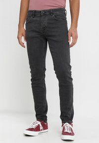 Hombre vestido con jeans ajustados de un negro desvanecido y zapatillas rojas de corte bajo con cordones blancos, de pie contra un fondo plano.