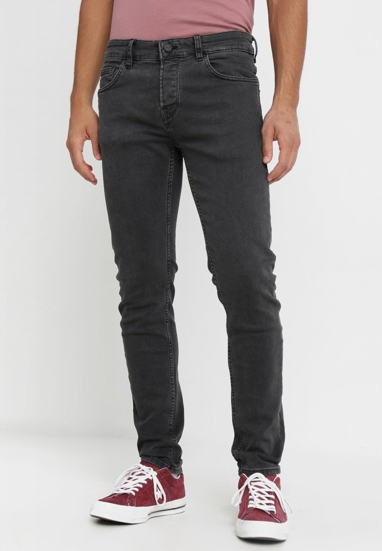 Hombre vestido con jeans ajustados de un negro desvanecido y zapatillas rojas de corte bajo con cordones blancos, de pie contra un fondo plano.