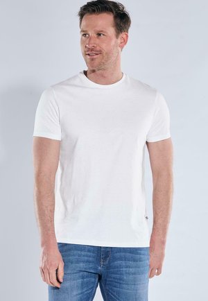 Man wearing plain white short-sleeve T-shirt and blue jeans, looking slightly left, standing against light gray background.