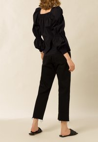 Frau, die eine schwarze Bluse mit Puffärmeln, schwarze Cropped-Jeans mit ausgefransten Säumen und schwarze Rutschsandalen trägt, steht vor einem beigen Hintergrund.