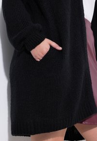 Černý pletený kardigan s ribovanouTexturou, který má dlouhé rukávy a boční kapsy. Lem je mírně delší vzadu.