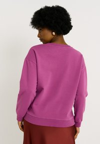 Mujer con cabello rizado oscuro que lleva un suéter morado de manga larga y una falda roja, de espaldas con pendientes de aro dorados visibles.