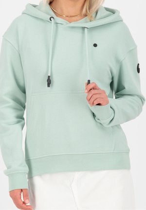 Femme portant un sweat à capuche vert clair avec des cordons, une poche avant et un pantalon blanc, tenant un cordon près de la poitrine.