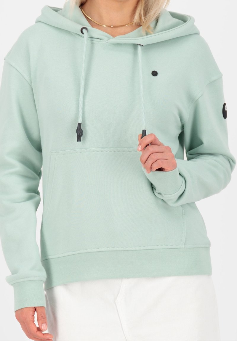 Femme portant un sweat à capuche vert clair avec des cordons, une poche avant et un pantalon blanc, tenant un cordon près de la poitrine.