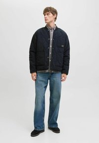 Marineblaue gewatteerde jas met drukknoopsluitingen en twee voorzakken, gedragen over een geruit shirt. Model draagt lichtblauwe jeans en zwarte schoenen.