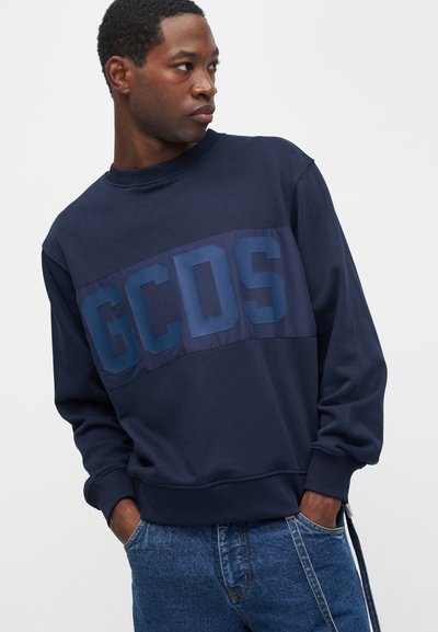 Marinblå sweatshirt med matt finish och glänsande "GCDS"-tryck över bröstet. Har ribbade muddärmar och en avslappnad passform, tillsammans med blå denim.