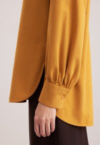 Senfgelb, langärmlige Bluse mit gerafften Bündchen, abgerundetem Saum und strukturiertem Stoff. Hand an der Seite des Kleidungsstücks positioniert.