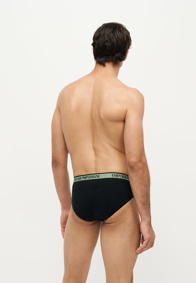 Homme vu de dos portant un slip noir avec une ceinture verte Emporio Armani sur un fond blanc uni.