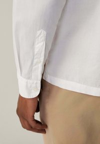 Camisa blanca de manga larga hecha de tejido liviano, con puños abotonados y dobladillo recto, combinada con pantalones color beige.