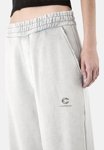 Gros plan sur une personne portant un pantalon de survêtement gris clair avec une ceinture élastique et le logo Camperlab près de la poche, la main reposant sur le côté.