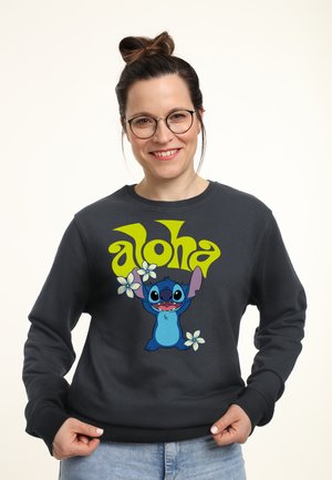 Vrouw met bril die glimlacht, draagt een donker sweatshirt met de blauwe cartoonfiguur Stitch en gele tekst "aloha" met witte bloemen.