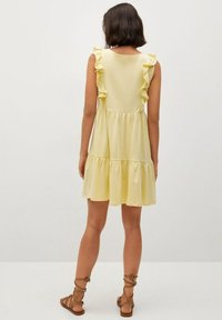 Žluté šaty bez rukávů s volánkovými detaily, vícestupňový design a měkká textura. Spárováno s hnědými sandály na řemínky.