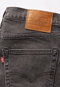 Jeans de mezclilla gris con un parche de cuero color marrón claro con marca en relieve y detalles, incluyendo costuras visibles y una etiqueta roja en el bolsillo trasero.